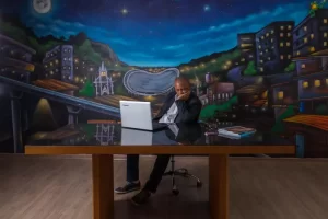 Celso Athayde que é o idealizador da Expo Favela, sentado em uma cadeira com o notebook em cima de uma mesa, atrás dele há um mural representando as casas das favelas.